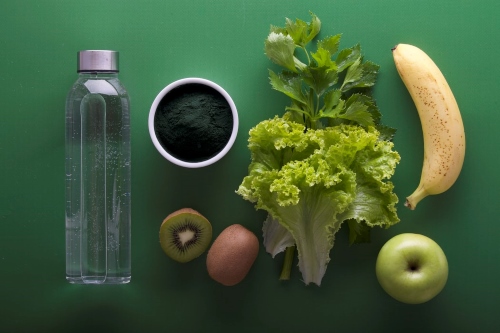 Water bottle, coffee, kiwi, egg, green fruit and vegetables laid out on green background