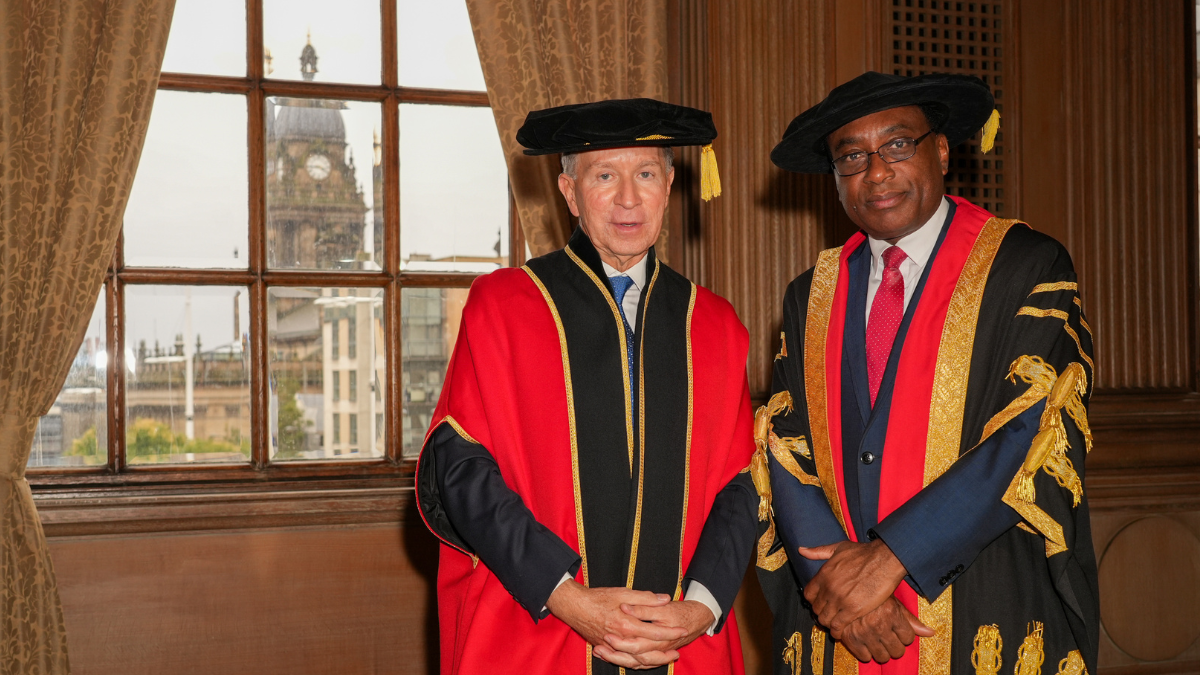 Leeds Trinity University's Chancellor, John Studzinski CBE, on the left, and Vice Chancellor, Professor Charles Egbu, on the right, posing at Leeds Civic Hall in red and black academic robes. Leeds Town Hall can be seen in the window behind them.
