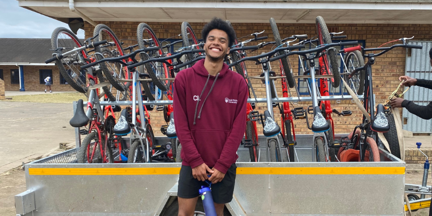 A male student wearing a burgundy hoodie smiles in front a bike rack