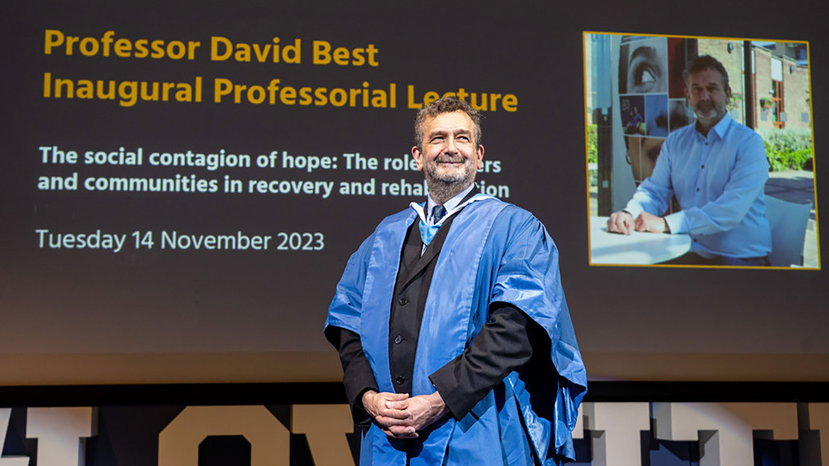 A male academic in a blue gown posing in front of a screen for his Inaugural Professorial Lecture.