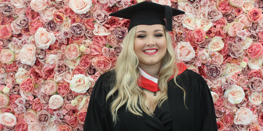 Blonde female wearing graduate cap stands in front of flower wall.