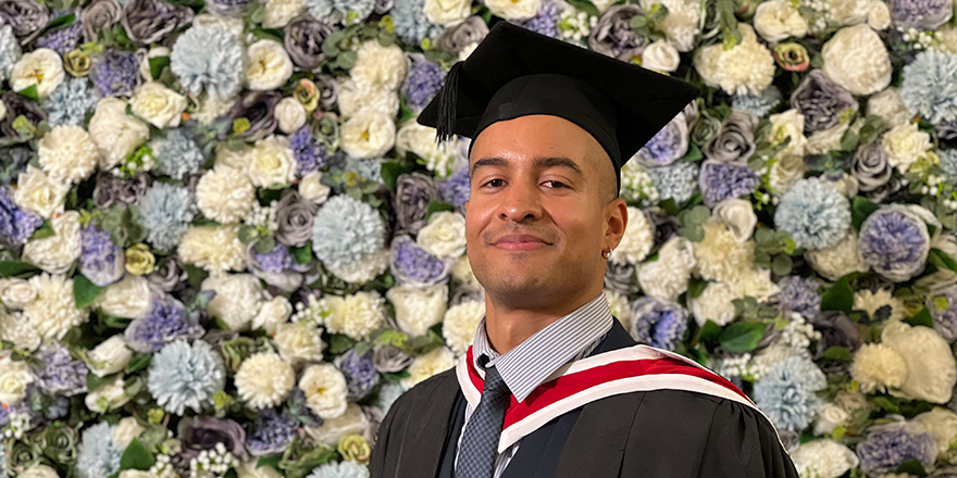 A mixed-race young man in graduation cap and gown posing in front of a wall covered in blue and white flowers.