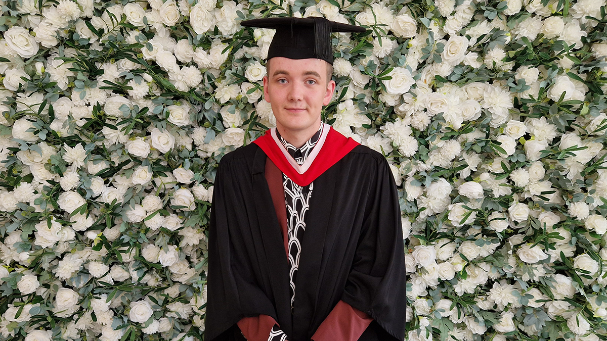 A student in graduation cap and gown smiling in front of a flower wall.