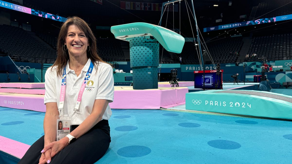 A woman with dark hair and a white top sits on a blue floor in front of gymnastics apparatus