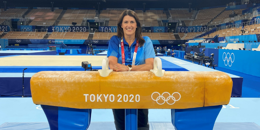 Woman with brown hair in tracksuit leans on pommel horse with Tokyo 2020 written on it.