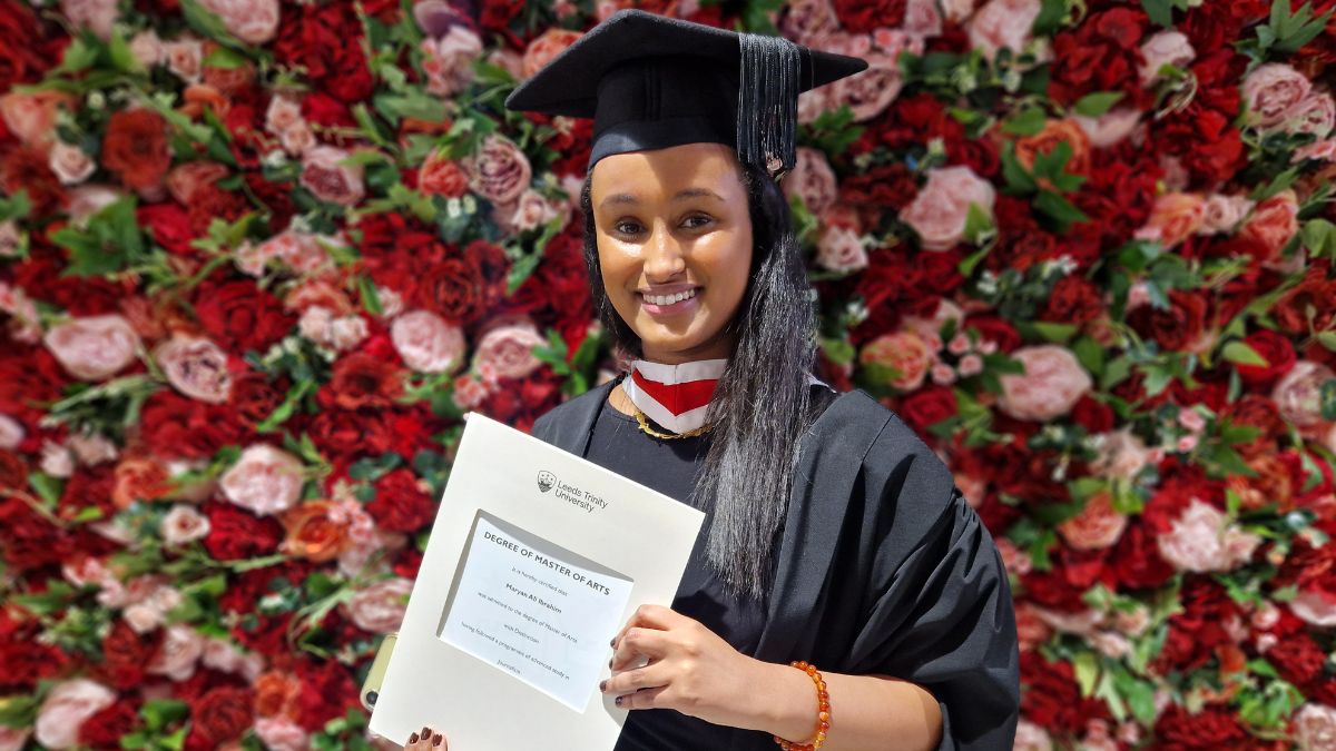 A girl in a graduation gown holds her certificate in front of a red and pink wall of flowers