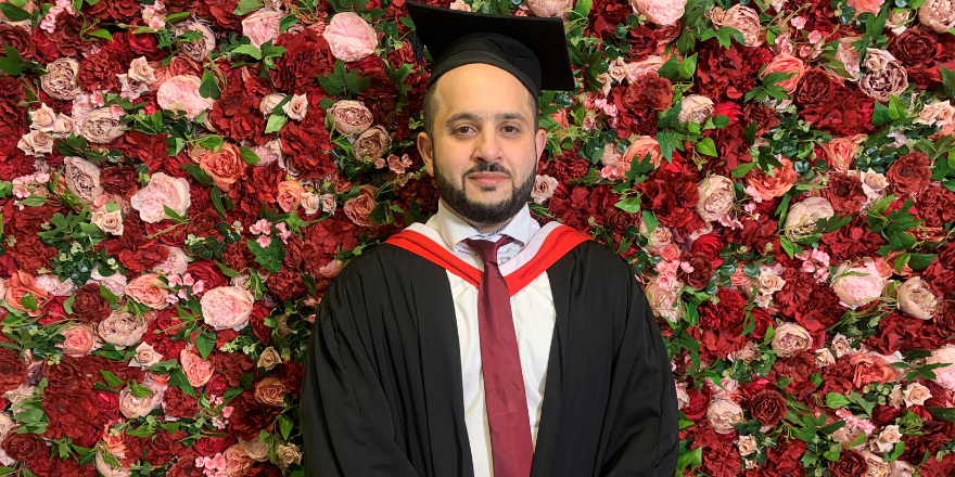 Male in graduation cap and gown stands in front of flower wall
