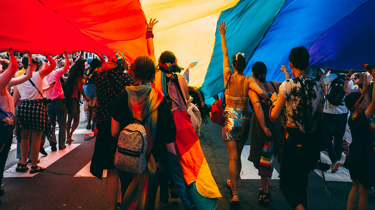 A group of people with their back turned to the camera, underneath a big rainbow flag and holding or wearing rainbow attire.