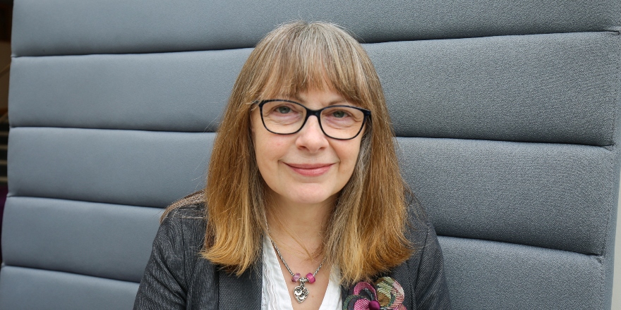 A headshot of a woman with brown hair and glasses, sitting on a grey sofa.