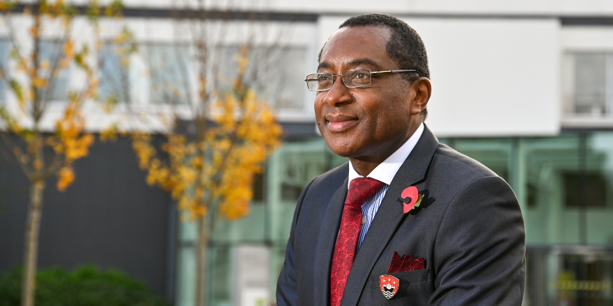Professor Charles Egbu sat in blue suit with red tie, yellow leaves on tree in background.