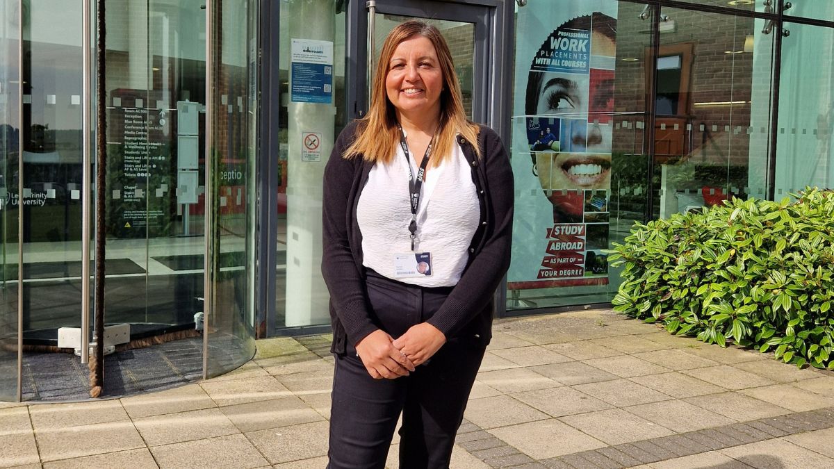 A woman wearing a white top a black cardigan and black trousers smiles for a picture in front of a building