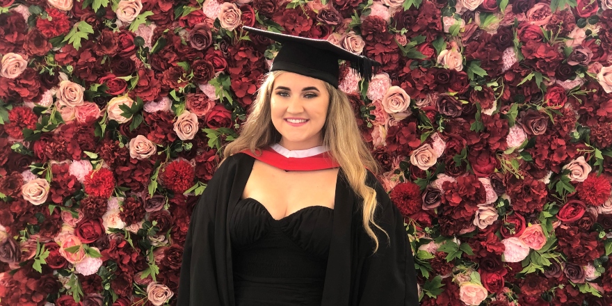 Female student in cap gown and black dress with red floral background.