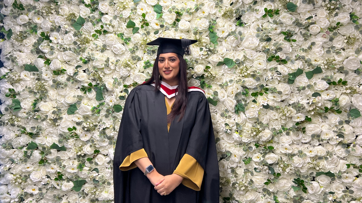 A student in graduation cap and gown smiling against a wall of white flowers.