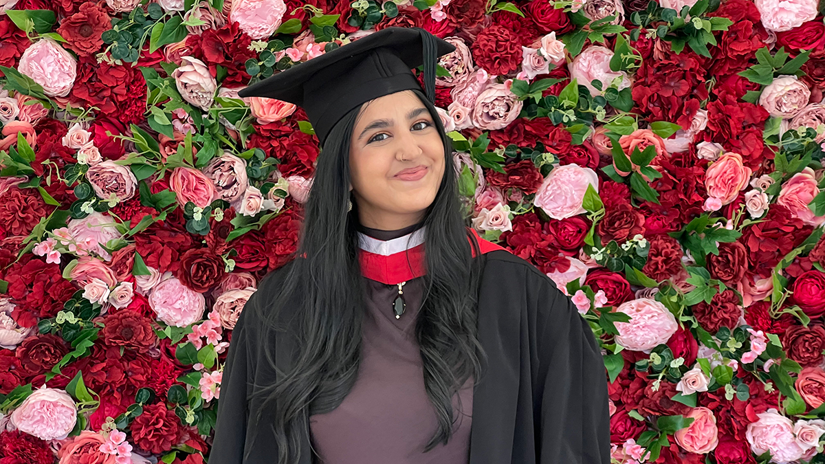 A student in cap and gown posing in front of a wall of red and pink roses.