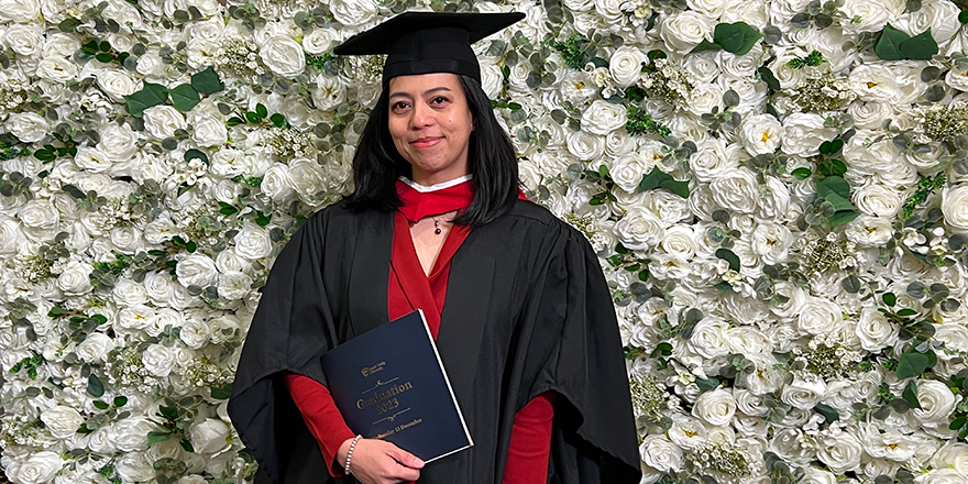 A Filipino woman in Graduation cap and gown posing in front of a wall covered in white flowers.